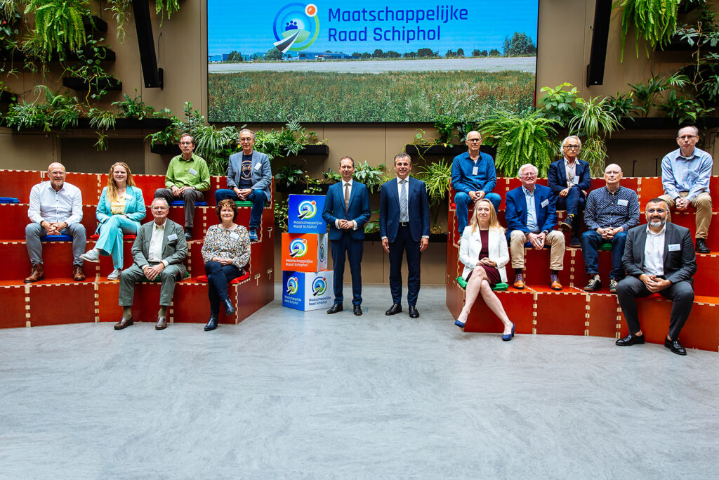 Groepsfoto van de leden van de Maatschappelijke Raad Schiphol, zittende op een houten tribune. In het midden staan voorzitter Eddy van Hijum en minister Mark Harbers. 