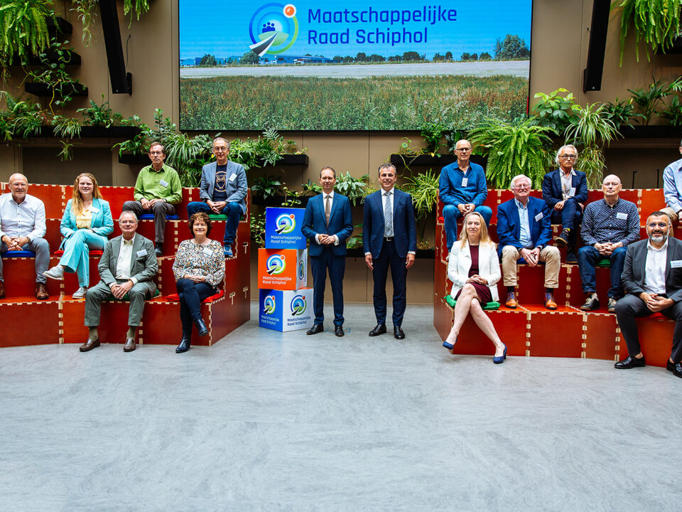 Groepsfoto van de leden van de Maatschappelijke Raad Schiphol, zittende op een houten tribune