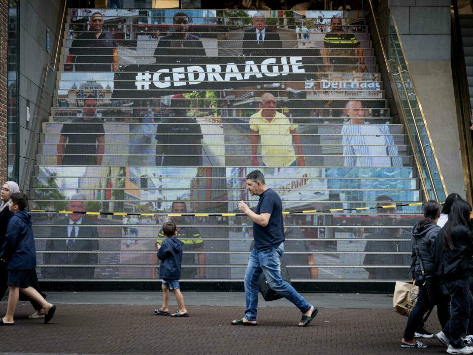 Spuimarkttrap met daarop de acht Haagse mannen in beeld, met in het midden campagneslogan #GEDRAAGJE